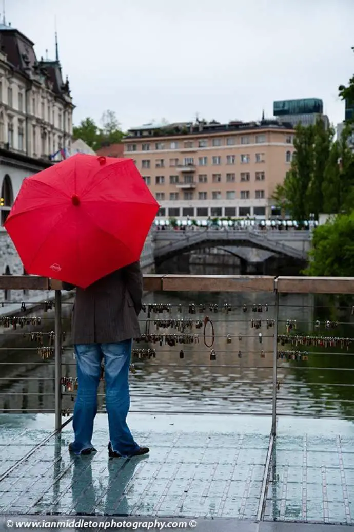 Man holding a red umbrella and standing on The Butchers’ Bridge looking over the Ljubljanica river towards the Trznica (market) Triple Bridge on a rainy day in Ljubljana, Slovenia. The wire fence on the bridge is covered with padlocks put there by locals and tourists. All this region was designed by Slovenia’s most celebrated architect, Jože Plečnik.
